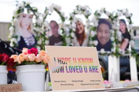 A memorial sign saying 'I hope u know how loved u are' next to a bucket of roses, with the faces of victims in the blurred background