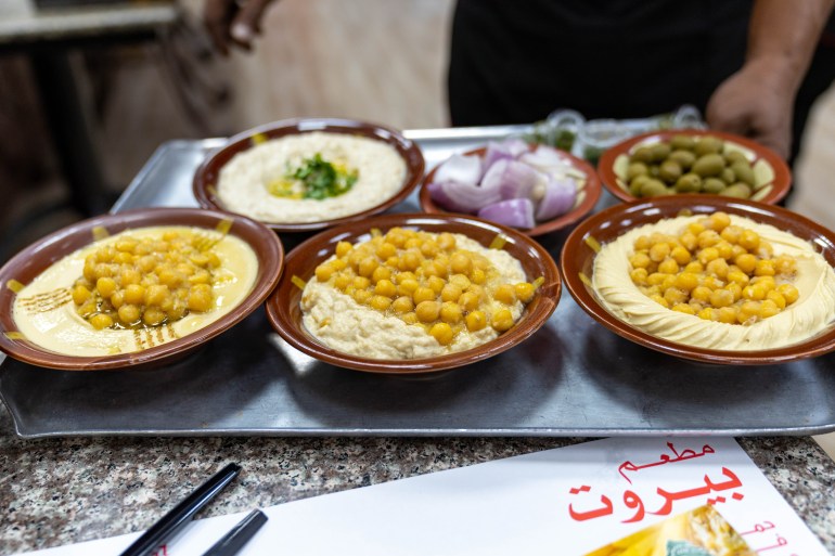 An aluminum tray with two bowls of hummus, a bowl of msabaha, a bowl of mutabal and some chopped onion and green olives
