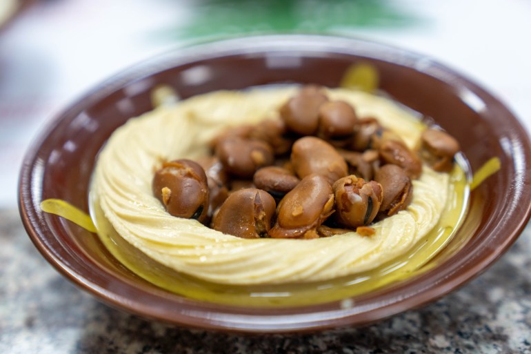 A brown bowl of hummust with swirled edges sits with a scoop of stewed fuul beans in the middle