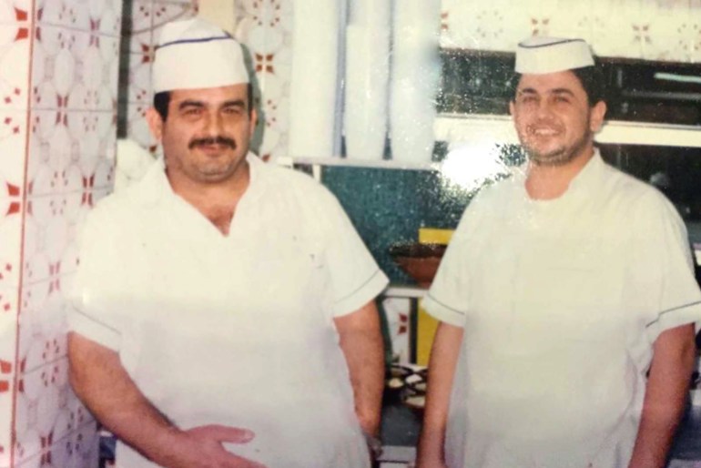 Two smiling men in a tiled kitchen, dressed in cook's hats and short-sleeved whites