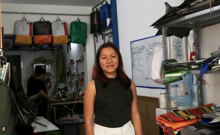 Kieu Anh Tran stands in her workshop with backpacks made from discarded tarpaulins behind her and offcuts on a metal shelf next to her. She looks content and there is someone working behind her.