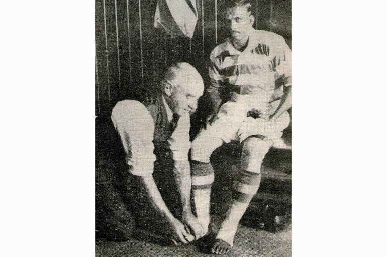 A photo of the footballer Mohammed Salim with his foot being wrapped in a bandaage by the Celtic FC trainer Jimmy McMenemy sitting on the floor.