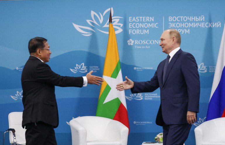 Min Aung Hlaing and Vladimir Putin shake hands at a summit in Vladivostok, Russia. They look pleased to see each other and are smiling. The flags of Myanmar and Russia are in the background.