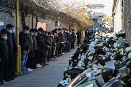 People dressed in black line up against a wall opposite a large pile of white flowers left by mourners. Their heads are bowed as they observe three minutes of silence for former Chinese President Jiang Zemin outside his old home in Yangzhou