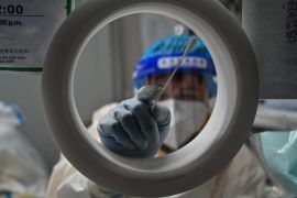 A health worker in a white hazmat suit pokes a swab towards the camera from the inside of a testing booth