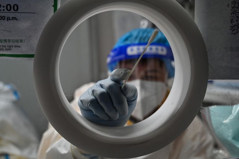 A health worker in a white hazmat suit pokes a swab towards the camera from the inside of a testing booth