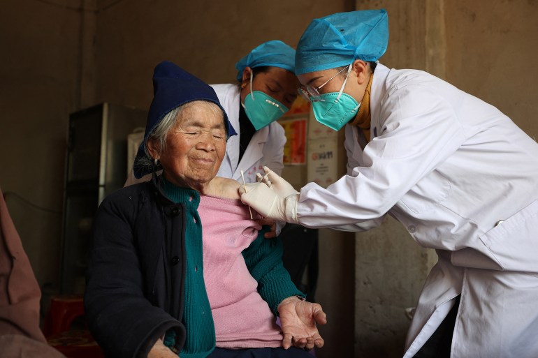 An elderly woman is given a dose of the COVID-19 vaccine from a healthcare worker. The woman looks relaxed. She's wearing a pink sweater, black coat and black beanie.