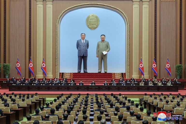 Giant statues of former Presidents Kim Il-Sung and Kim Jong Il stand at the front of the hall under which is a long table that leaders are seated behind. The rows of attendees seats face the table and statues