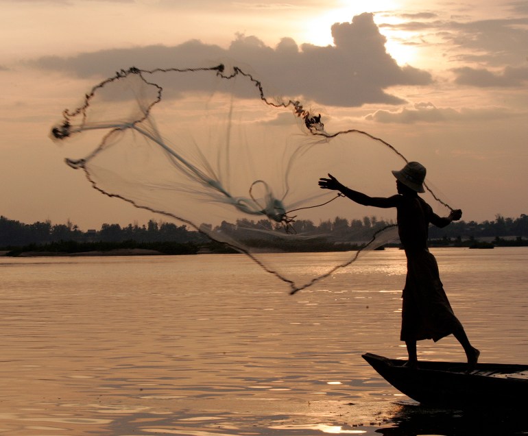 A fisherman throws his net out on the Mekong. He is silhouetted against a dawn sky and is standing at the bow of his small boat