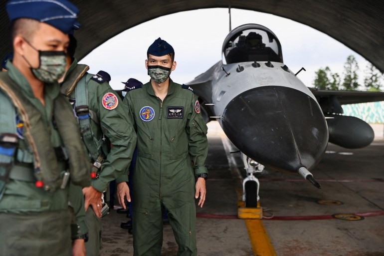 Two air force officers in combat uniform and wearing face masks standing in a hangar at an airforce base in the Penghu Islands. There is a military aircraft to one side of them