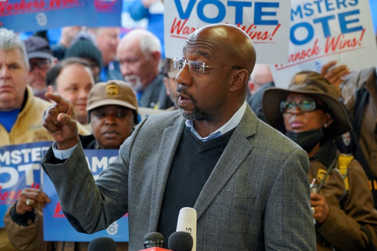 Raphael Warnock speaking at a campaign rally with supporters behind him holding placards.