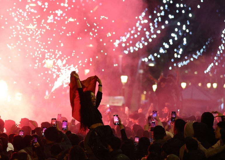 Fan sits on shoulders of someone in the crowd and holds up red flag in joy. Many in the crowd are holding up their mobile phones to film the celebrations. Everything is pink and there look to be firecrackers or flares above the crowd