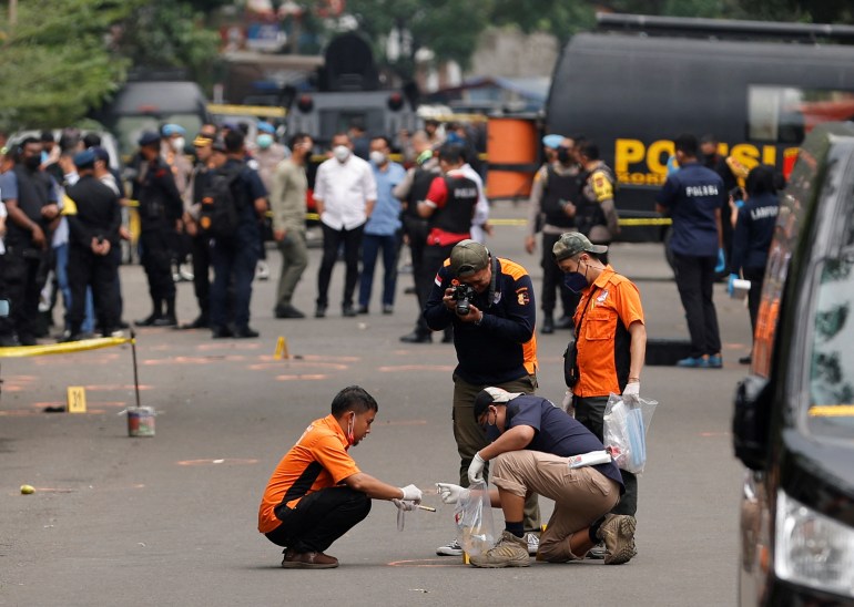 Officers in orange shirts inspect the scene of the Bandung suicide bombing. They are kneeling down on the road and putting evidence into a clear plastic bag. There is a crowd of onlookers behind with the blast area cordoned off by yellow police tape
