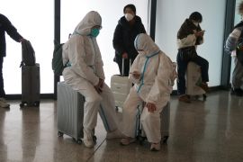 Travellers in full white hazmat suits sit on their luggage as they wait to board a train. One is looking at their phone