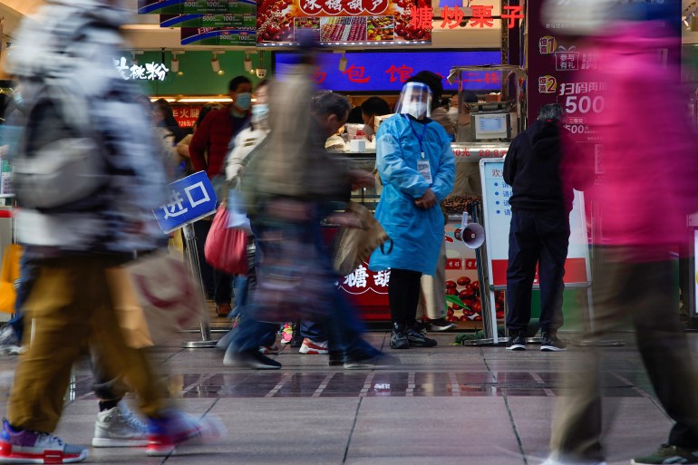 People walking on the street outside a food shop where a worker outside is wearing a blue disposable medical gown, face mask and transparent plastic face shield.