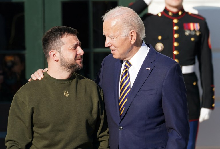 Biden has his arm around Zelenskyy, who is looking up at him with an emotional expression on his face. Zelenskyy is wearing a dark green sweater with a small gold printer symbol of Ukraine's armed forces at the top centre 