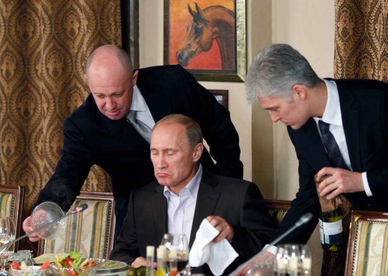 FILE - In this Friday, Nov. 11, 2011 file photo, businessman Yevgeny Prigozhin, left, serves food to Russian Prime Minister Vladimir Putin, center, during dinner at Prigozhin's restaurant outside Moscow, Russia. Russian news outlets say thousands of Russians have been deployed in Syria by a shadowy, private military contractor linked to Prigozhin. When President Vladimir Putin announced Monday Dec. 11, 2017 that Russia’s campaign in Syria was drawing to a close, he did not mention this secret force. (AP Photo/Misha Japaridze, Pool, File)