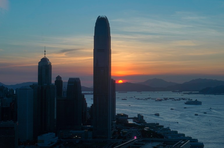 Hong Kong's city bay in the fading light, with towers stretching up to a sky coloured by blue, orange and yellow and in the background, a sun halfway behind a mountain range that sits at the back of the sea