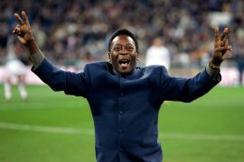 Pele running towards the camera on the field, wearing a navy, buttoned-up suit, and holding both his arms up at this sides, doing the peace sign