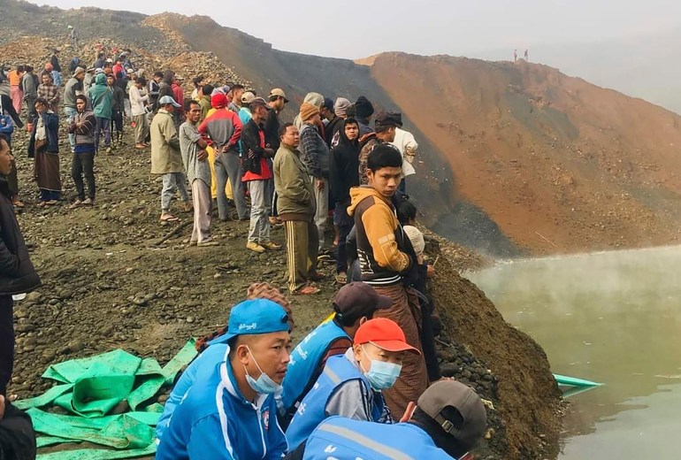 People standing around the rim of a lake formed by gold mining in Hpakant after a landslide. Many look cold and worried.