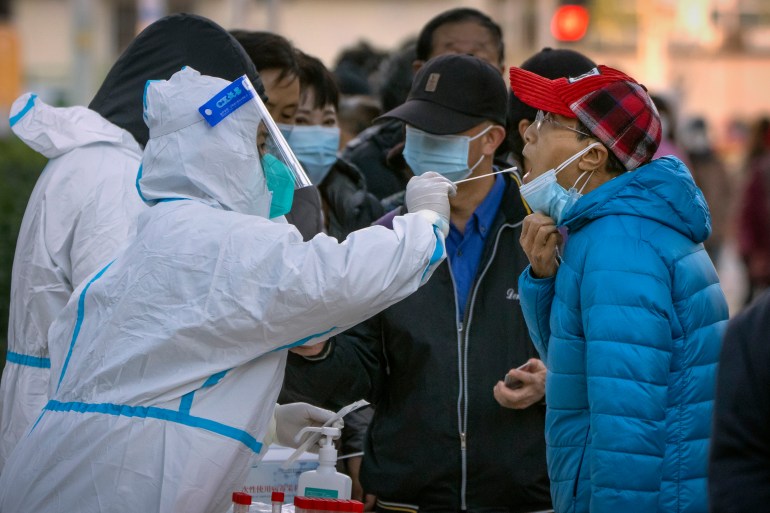 Woman in full protective gear holds a swap next to a man's open mouth. Others wait behind with masks on..