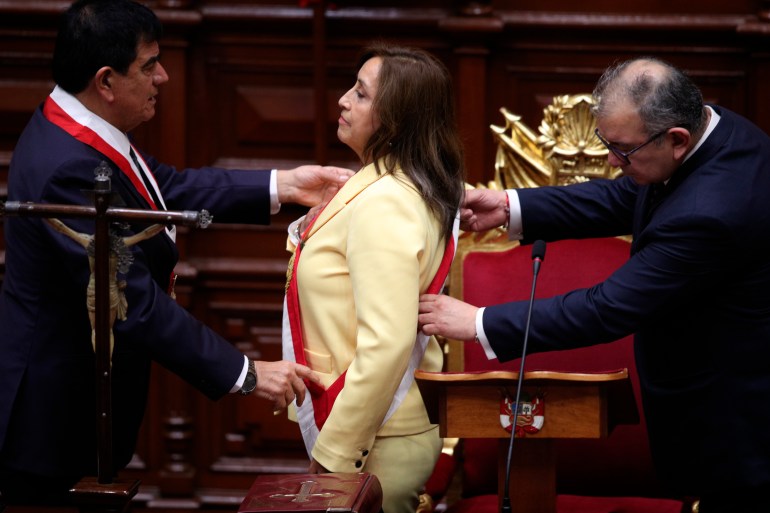 Boluarte stands in pale yellow suit two men, on either side, fix a red and white sash over her shoulder