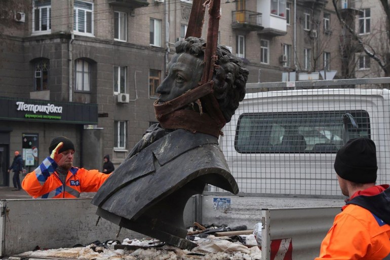 Alexander Pushkin's stone bust us being lowered onto the back of a truck by what is likely a crane that is cut off from the top of the photo. Two workers in orange gear stand on either side of the truck. Two workers 