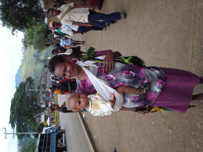 Mother is wearing a purple dress. Her child is on her hip, held with a white strap tied behind her neck