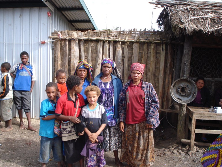 Three mothers with four children stand against the backdrop of a wooden fence and a hut with a straw roof. Two more children lean against what looks to be a corrugated iron shed on the other side of the fence