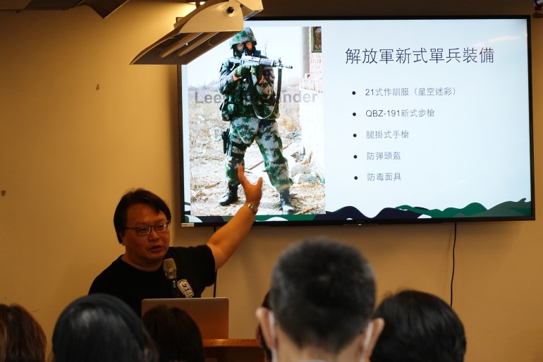 Cheng Hui-Ho giving a presentation to members of the public about the equipment carried by PLA soldiers. There's a slide on the wall showing a soldier with his gun lifted and writing in Chinese characters on the right hand side. Cheng is pointing at the slide, which is behind him