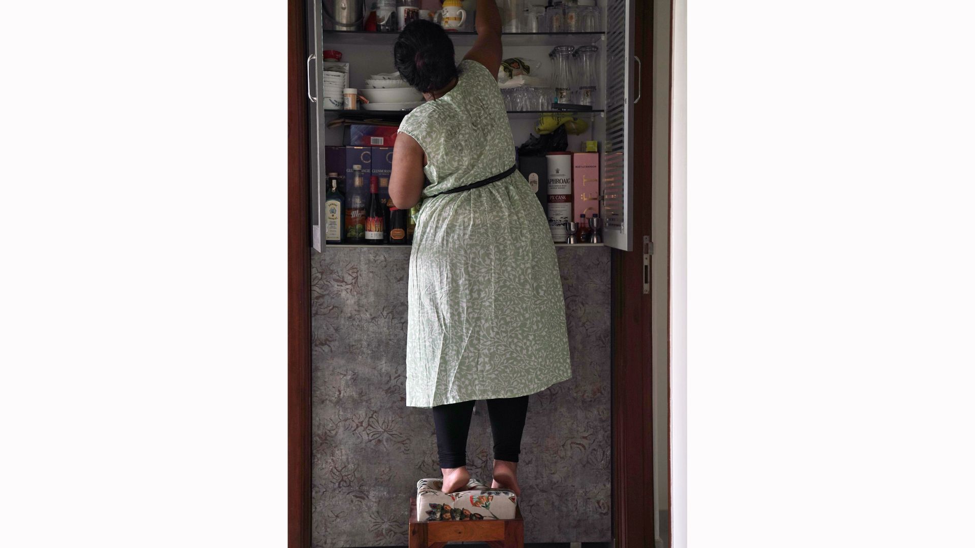 Akanksha stands on a stool in front of her pantry cupboard, lifting onto her toes to reach something in the top shelf