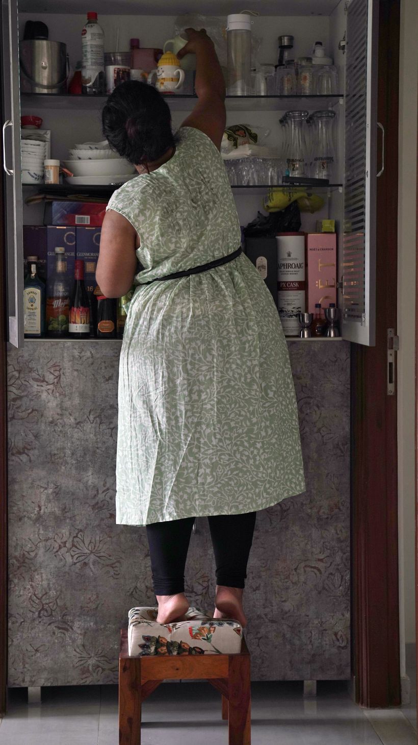 Akanksha stands on a stool in front of her pantry cupboard, lifting onto her toes to reach something in the top shelf