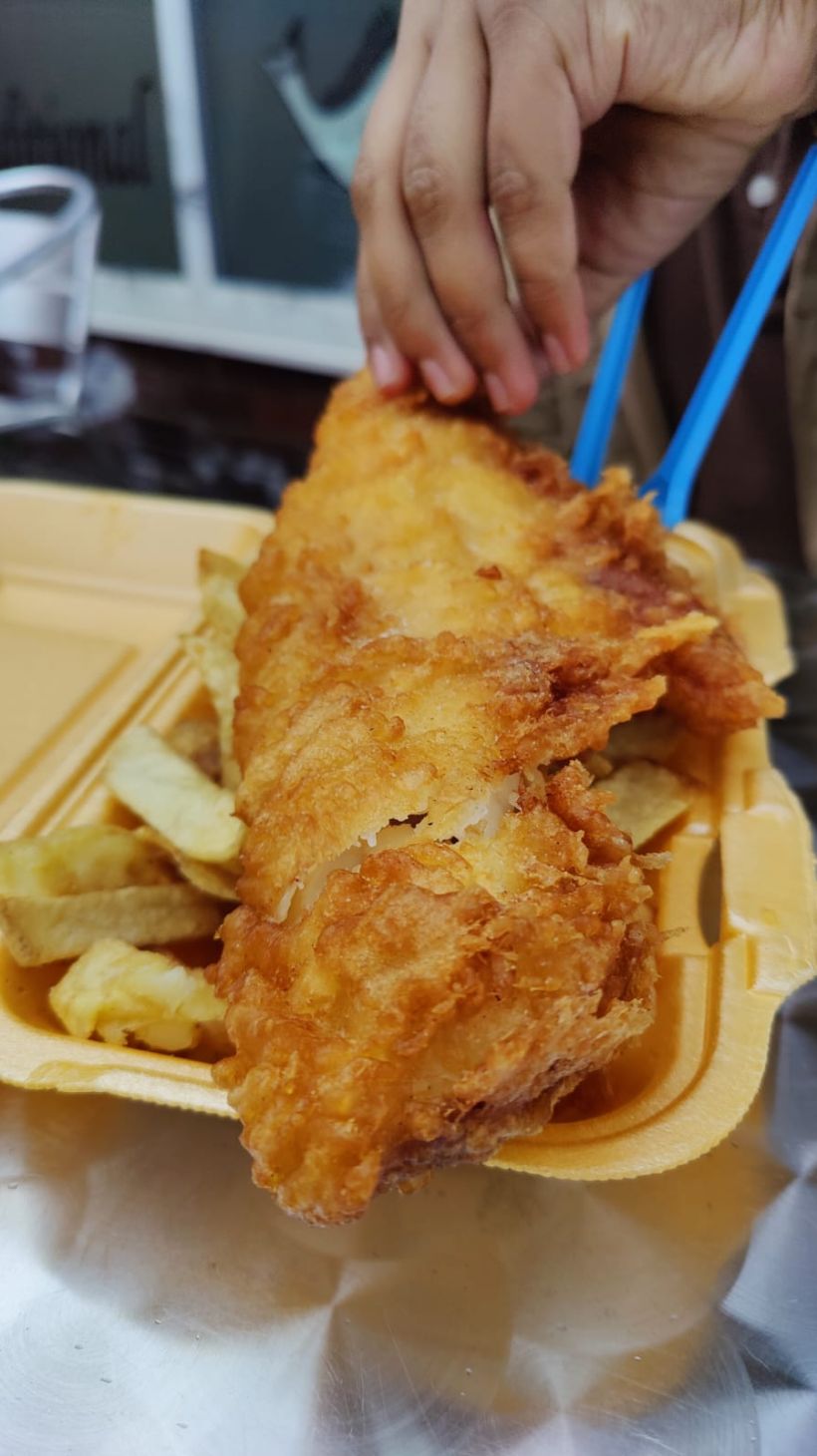 A closeup of a takeout container of fish and chips with Boba's hand grabbing the battered fish to start eating