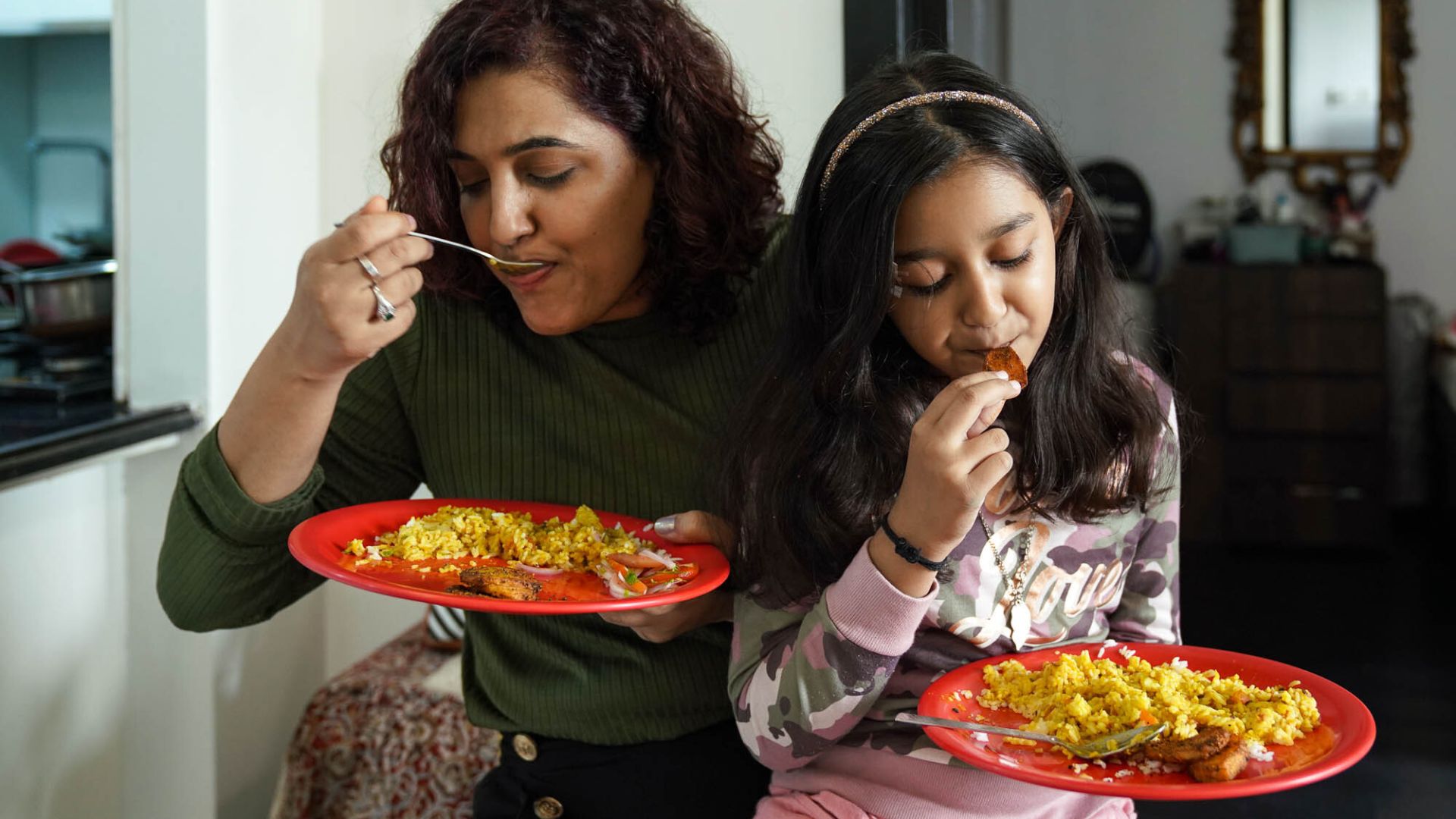 Khushboo sits with her daughter on her lap, each eating their meal out of a red plate