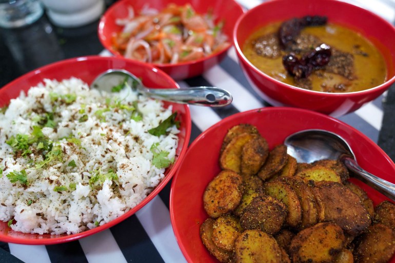 A meal set out, rice with herbs, fried potatoes, dal, and salad, each in a red bowl