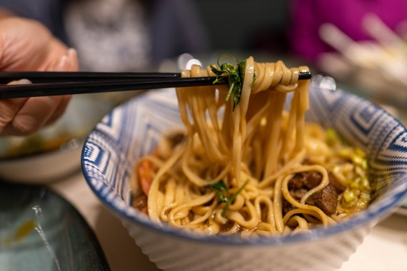 A hand holds a bite of noodles up with a pair of chopsticks out of a bowl of beef broth noodles