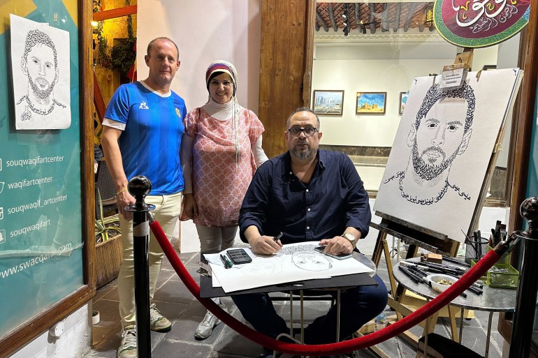 Khaled Almesawy sits with a sketch resting on his knees. There is a sketch of Messi on an easel to his left and another hanging on the wall to his right. Two people pose for the photo behind him.