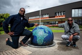 Jacques and Mahmoud squat next to a sculpture in the shape of the world outside the Mangere Refugee Resettlement Centre. They look amused