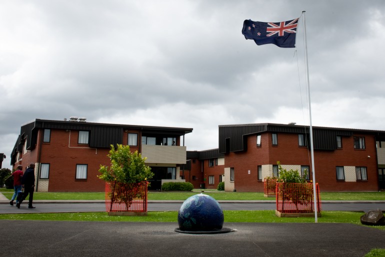 Mangere Refugee Resettlement Centre in Auckland, New Zealand. It is a two storey red brick building with a brown roof and there are balconies. A New Zealand flag is flying and the garden is neat and tidy. Two people are walking towards the building carrying shopping bags