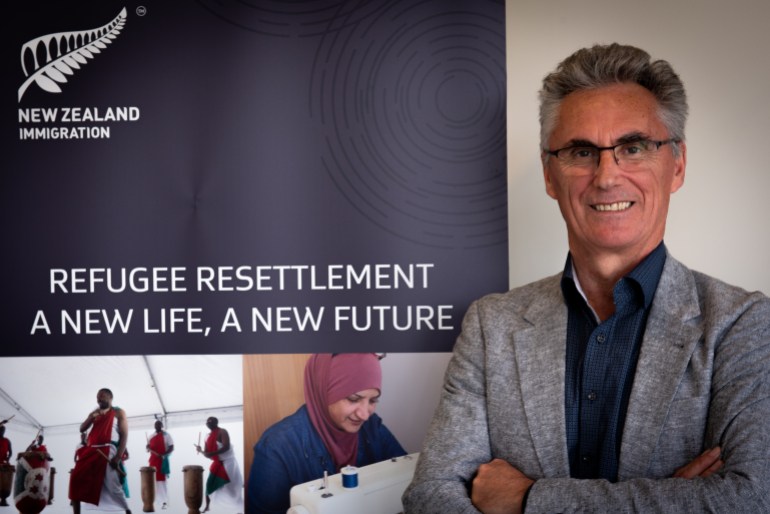 A formal portrait of Qemajl Murati, manager of Refugee Quota Program for Immigration New Zealand, He is wearing a light grey jacket and blue open-necked shirt and is smiling