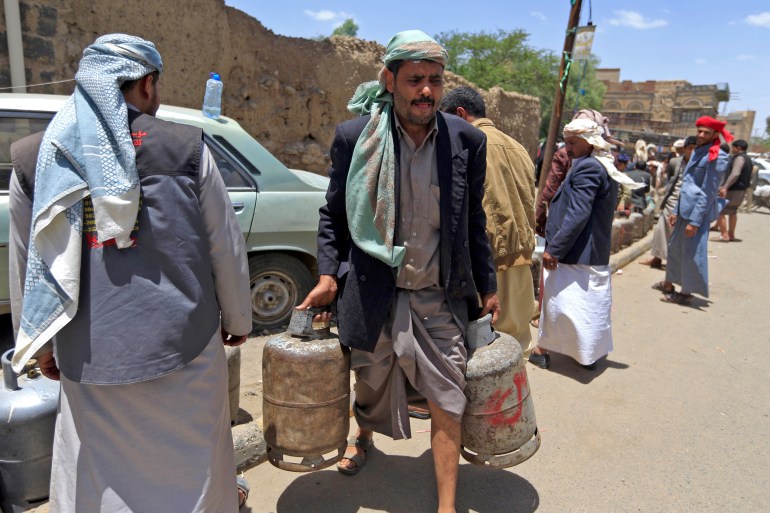 A photo of a man carrying two gas cylinders with people and a row of gas cylinders in the background.