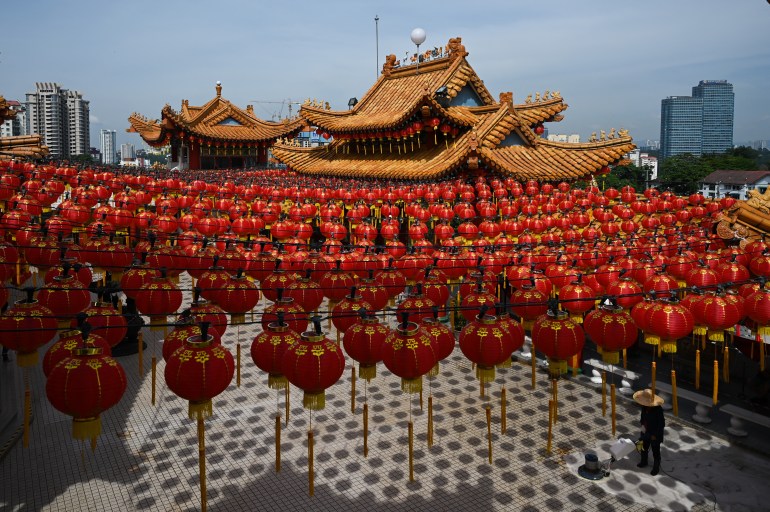 A view of the Thean Hou Temple in Kuala Lumpur. It has an orange roof and there are red lanterns hung across the courtyard. A man is sweeping the floor.