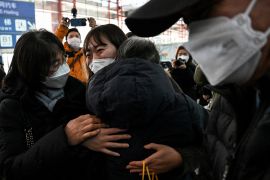 A woman in a face mask ehugs two other people at the international arrivals area of Beijing airport, She looks emotional. A man in a face mask is walking into the picture from the right. Everyone is wearing thick winter coats