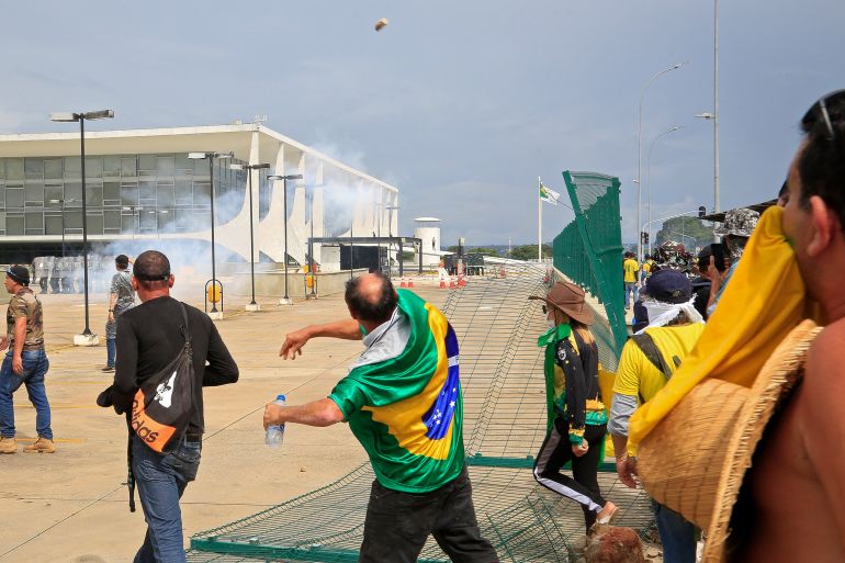 A man with a Brazilian flag around his shoulders throws something towards riot police in Brasilia, Brazil, as Bolsonaro supporters storm key institutions. There is tear gas smoke around them.