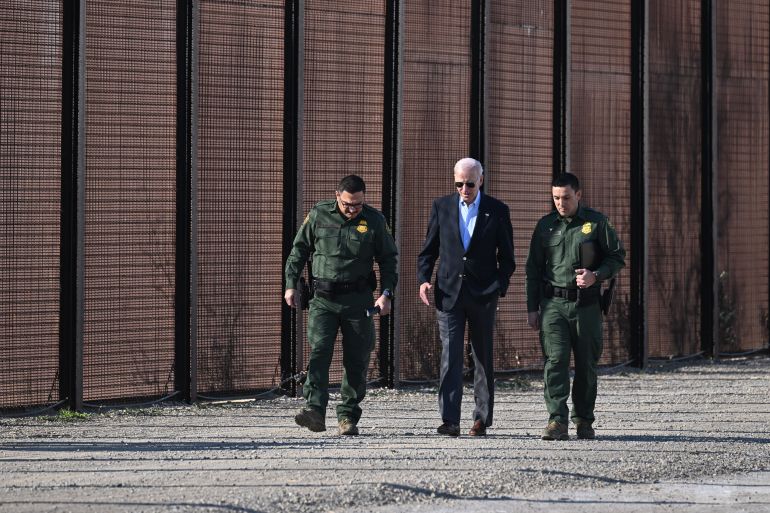US President Joe Biden in a dark blue suit and sunglasses walks along the US-Mexico border with uniformed US officials. There is a large wall behind them.