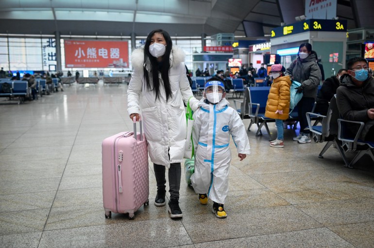 Woman is wearing a long white coat, a mask and pulling a pink suitcase. The child next to her is clad in PPE, a see-through face shield and is wearing a mask. Other travellers sit on chairs to the side, all wearing masks and coats or warm jackets.