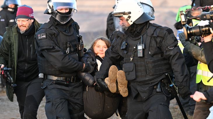 Greta Thunberg being carried away by two German police officers. She is not struggling and looks relaxed. The police are wearing white helmets and black uniforms