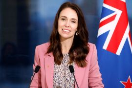 Ardern in a pink jacket smiles from behind two microphones with a New Zealand flag behind her