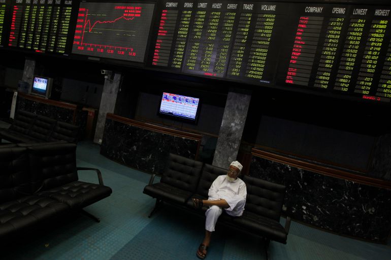 A trader sits on a couch, underneath an electronic board displaying share prices