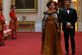 Jacinda Ardern with her partner walks through Buckingham Palace wearing a brown silk dress and a traditional Maori cloak known as a kaxx, They are both smiling. A beefeater is standing to the left and there are paintings and antiques behind them